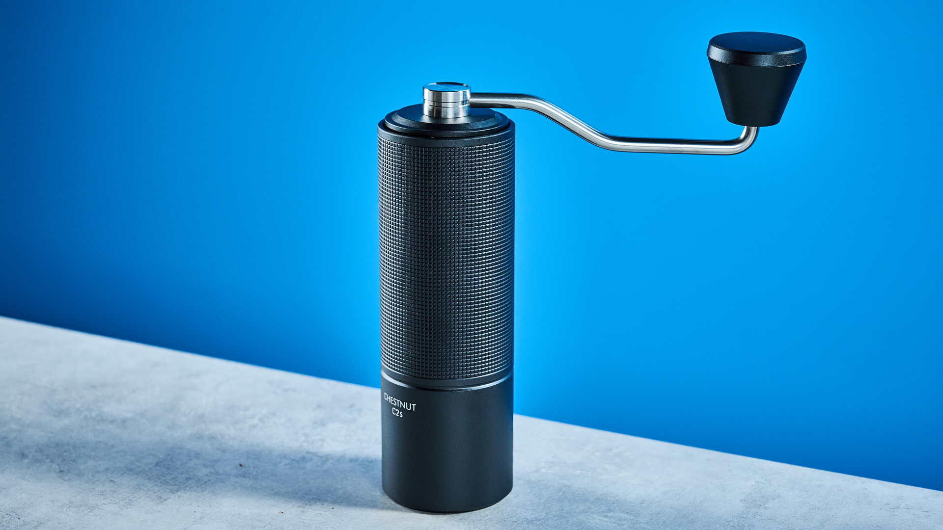 מטחנת הקפה של timemore chestnut c2s בשחור, מציגה את ארכובה היד ואת תא התפיסה שלה שצולמו על רקע המדריך הכחול של Tom's