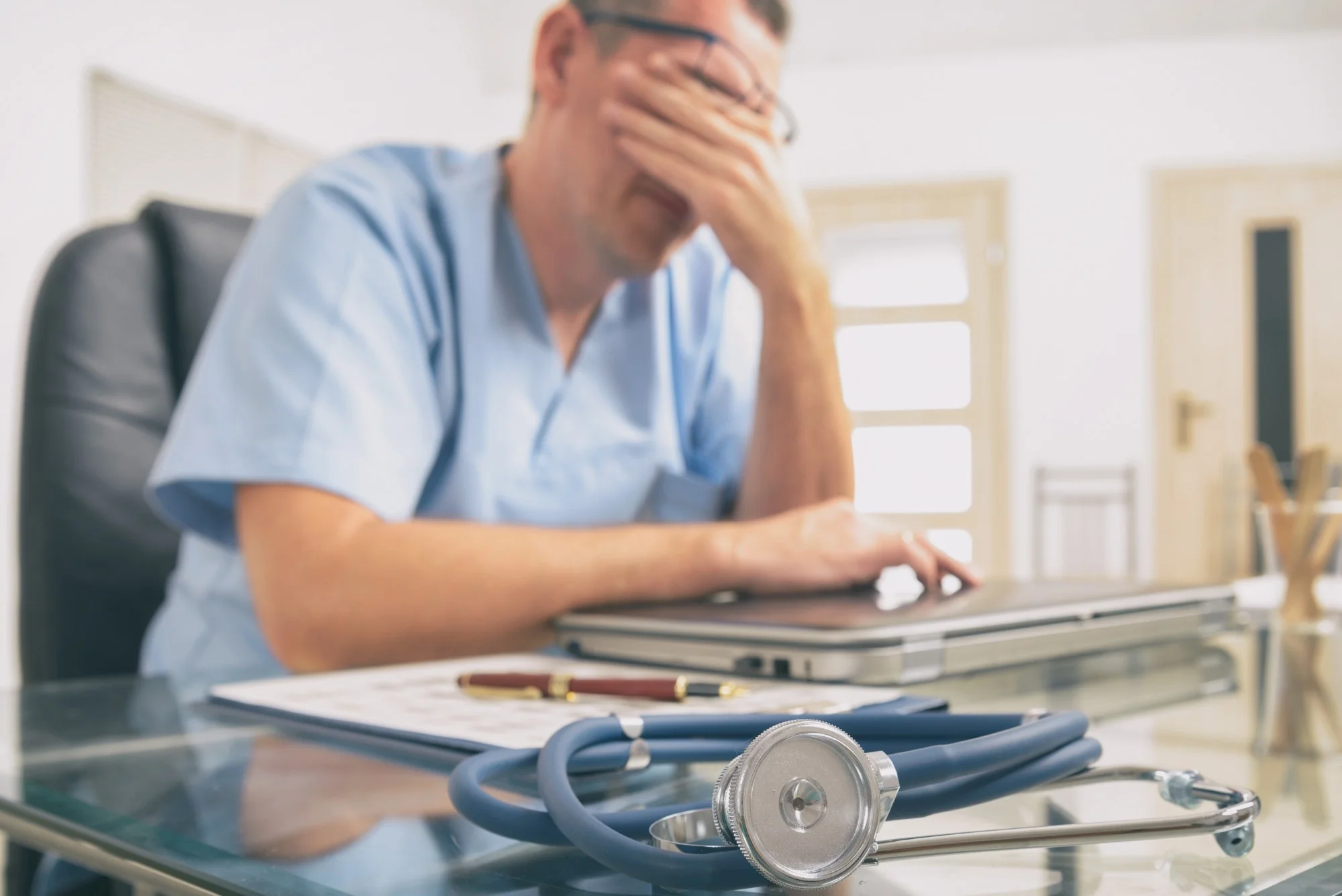 Overworked doctor sitting in his office with head in his hands. Focus on stethoscope in foreground