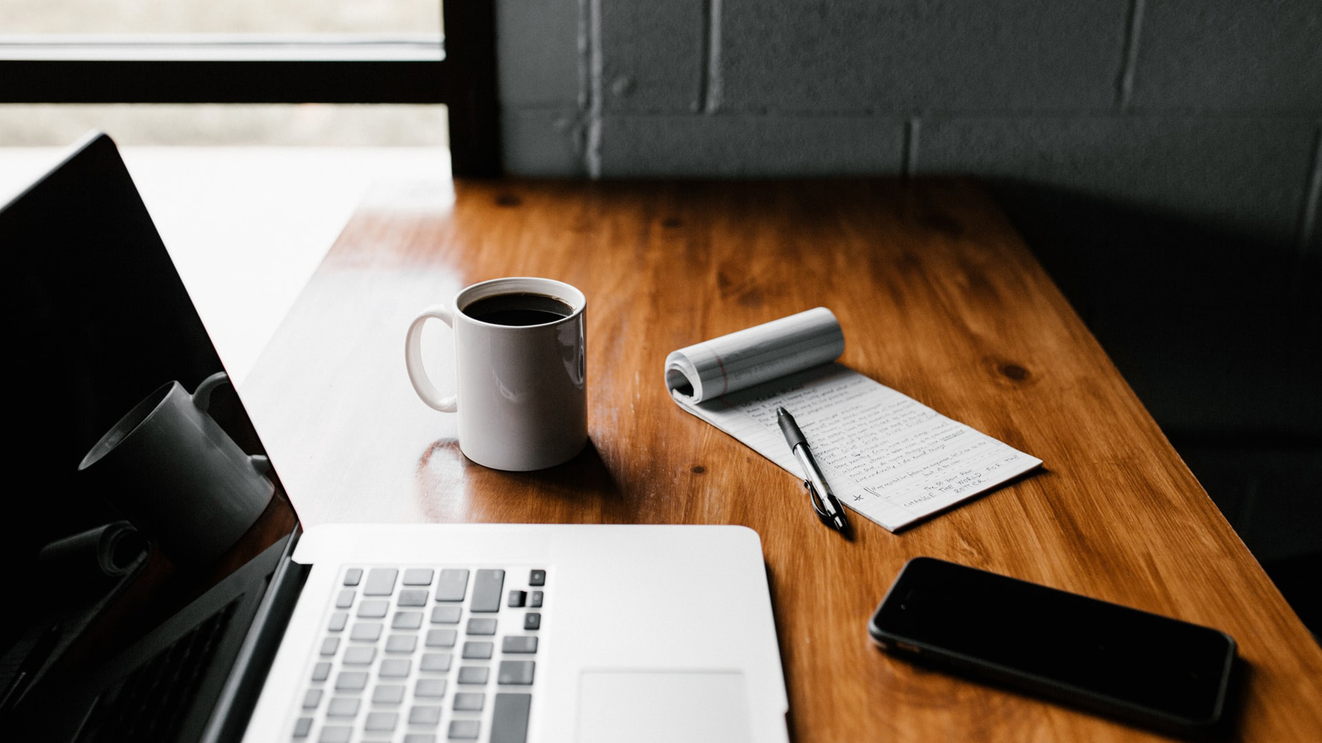 מחשב נייד פתוח על שולחן העבודה ליד פנקס רשימות ואייפון