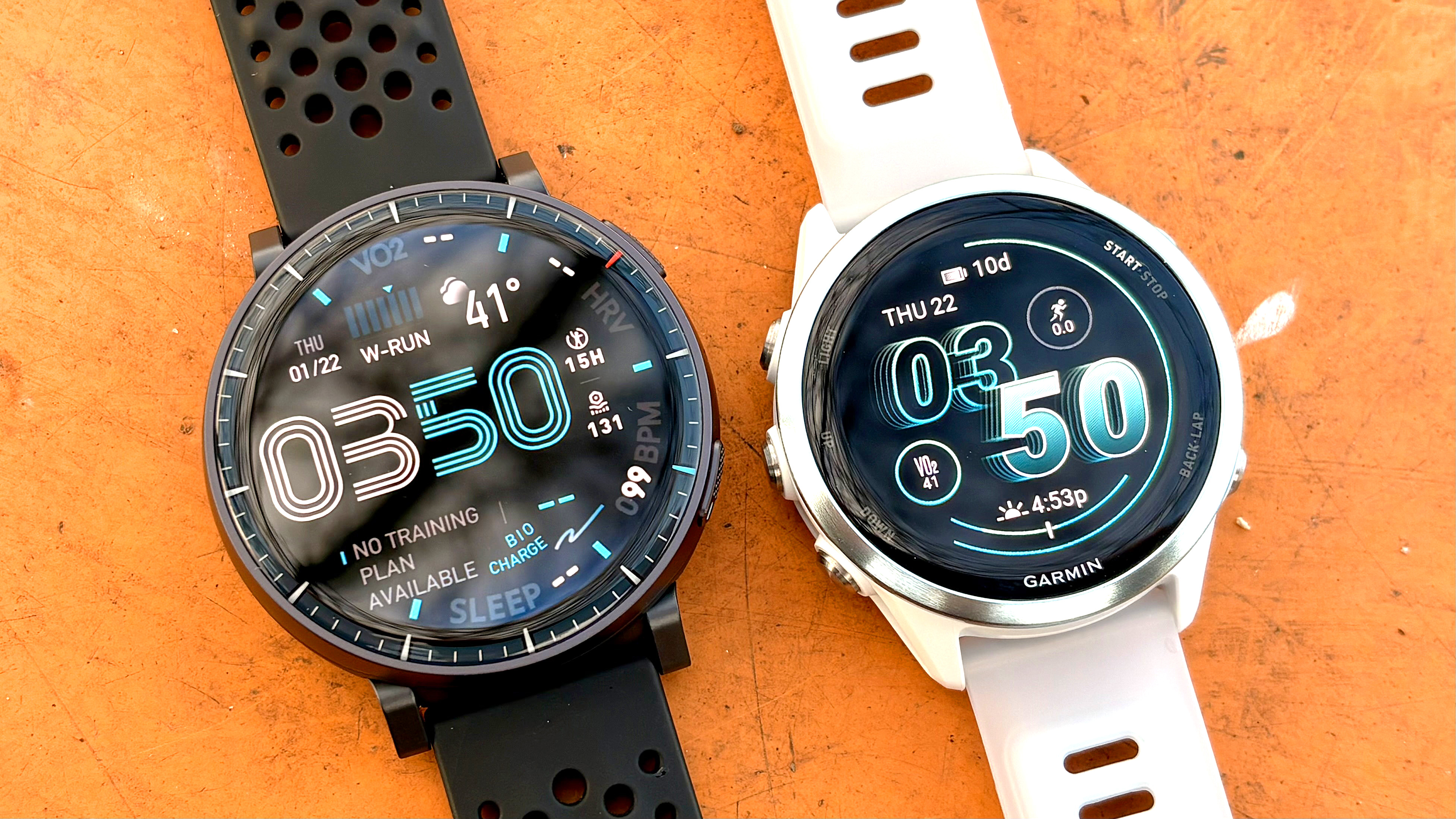 תקריב של ה-Amazfit Active Max ליד ה-Garmin Forerunner 570 על שולחן כתום בוהק