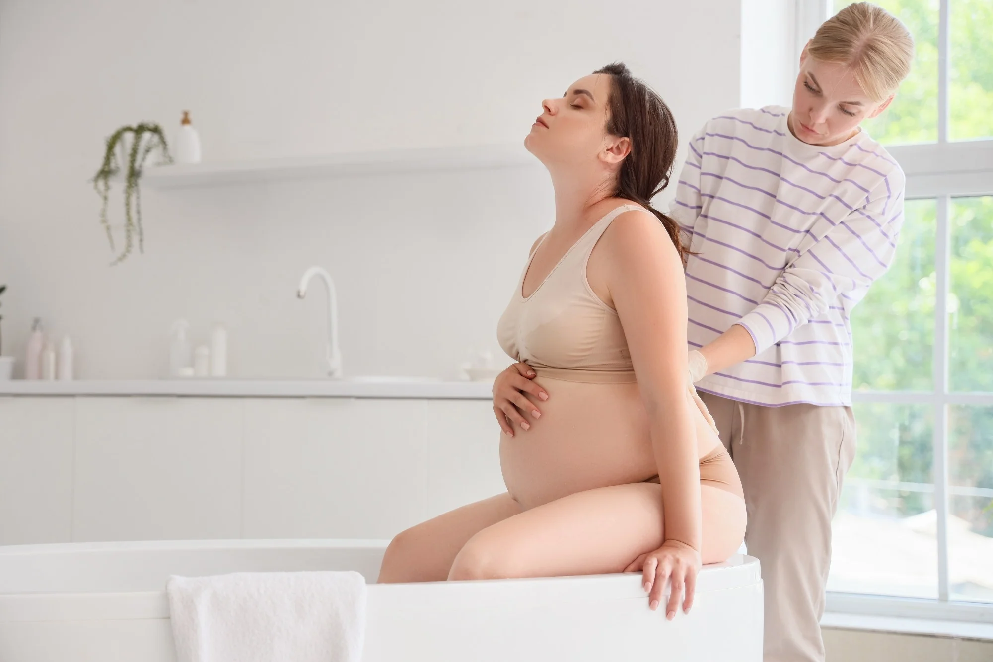 Young doula and pregnant woman sitting in bathtub at home