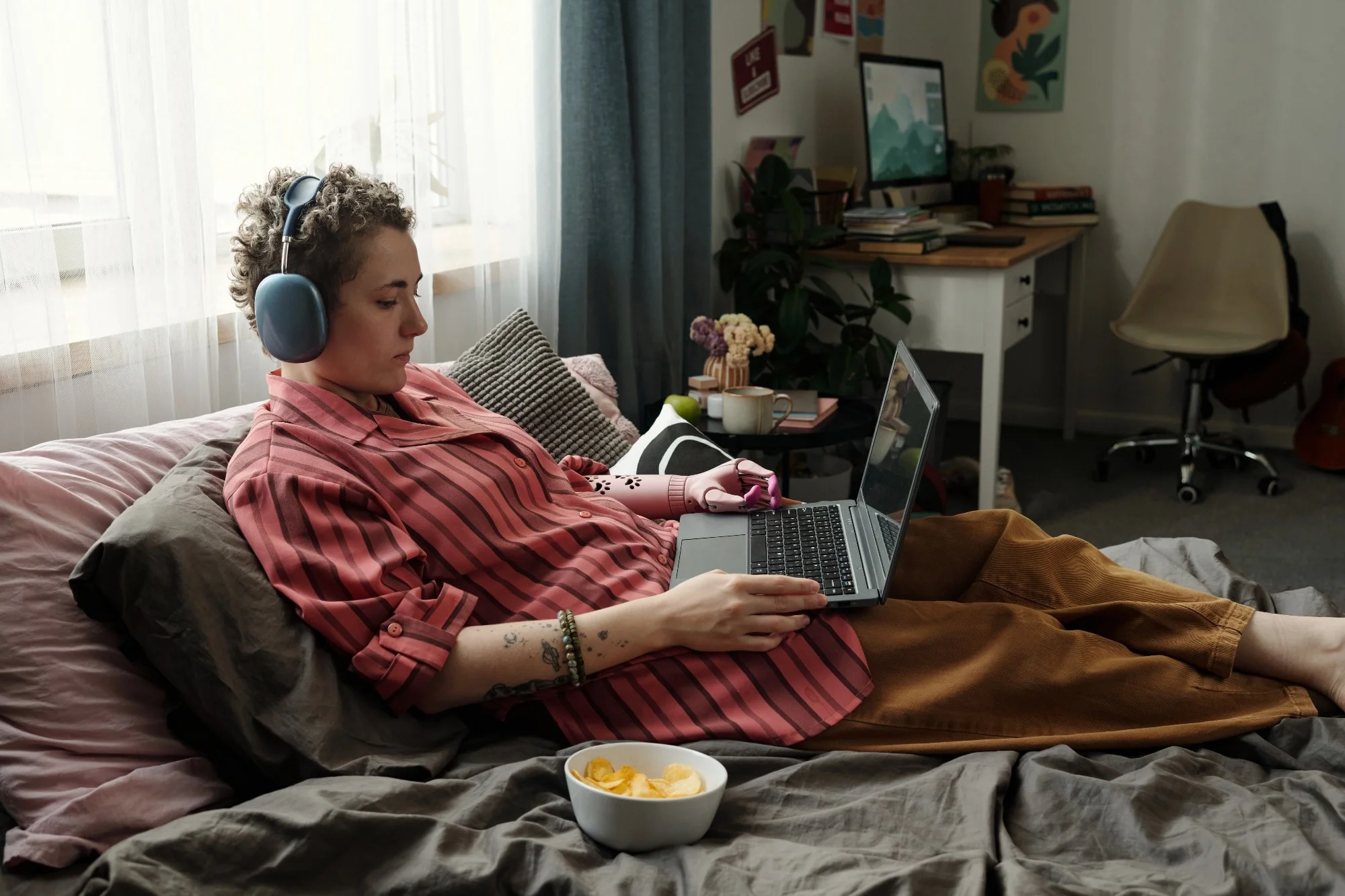 Caucasian young adult woman lying on bed using laptop and wearing headphones, holding smartphone in hand, surrounded by snacks, demonstrating signs of gadget addiction in bedroom