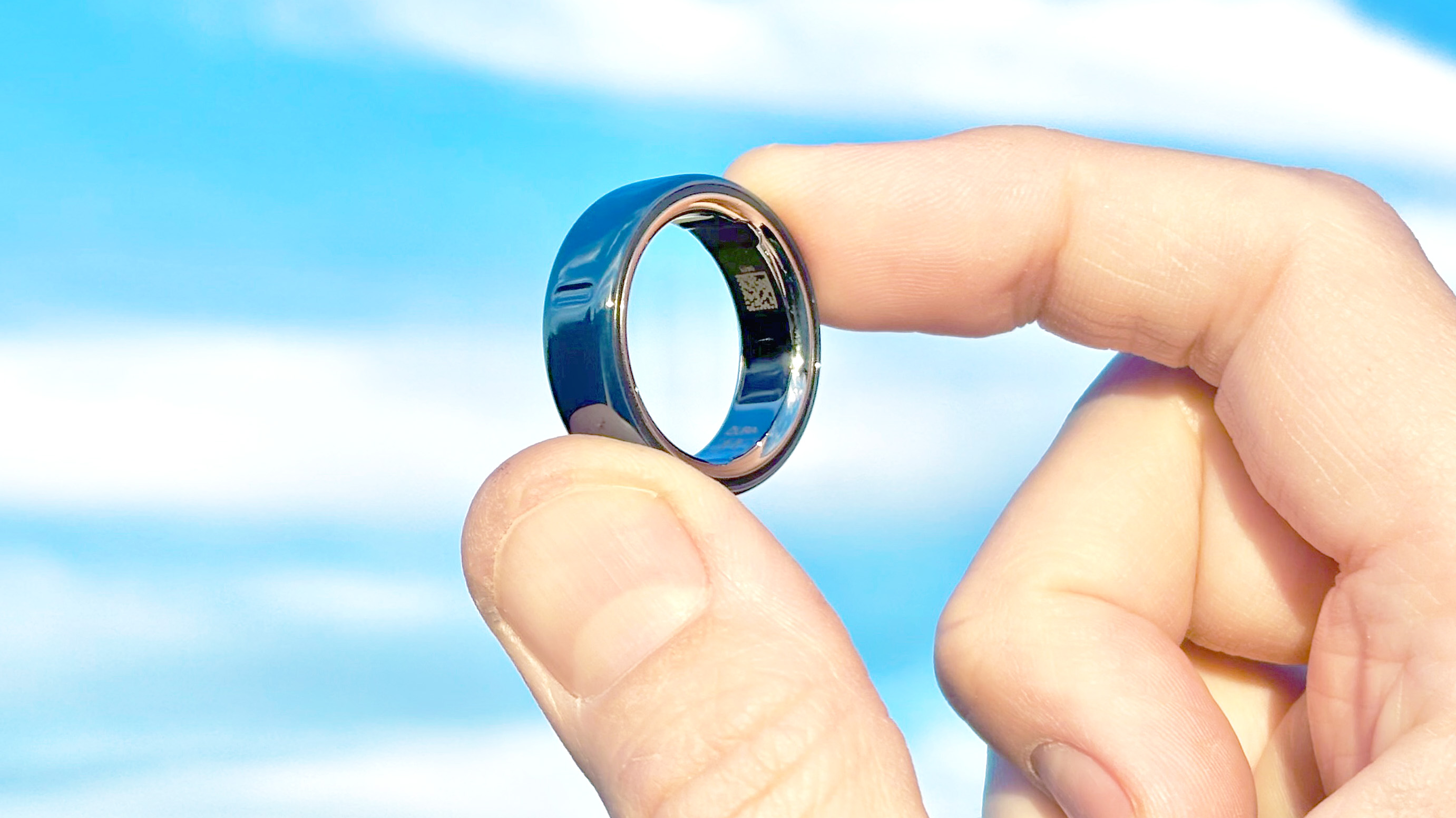תקריב של קרמיקה Oura Ring 4 זורחת באור השמש ביד משתמש עם שמיים כחולים ועננים מאחוריה