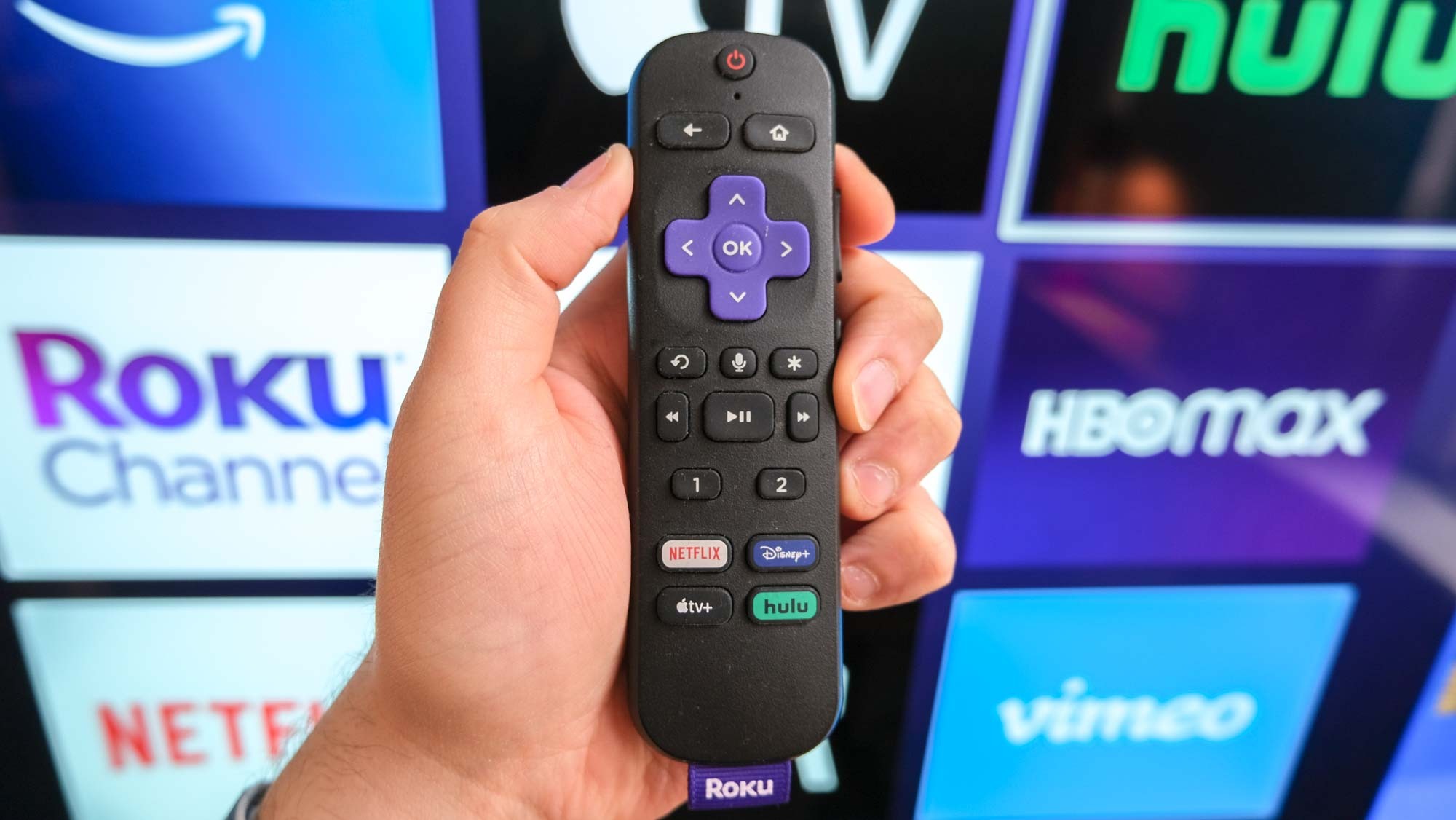 Roku Voice Remote Pro ביד מול טלוויזיה המציגה את מסך הבית של Roku.