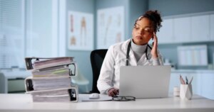 Exhausted Hospital Doctor Surrounded By Mountain Of Paperwork And Calculator.