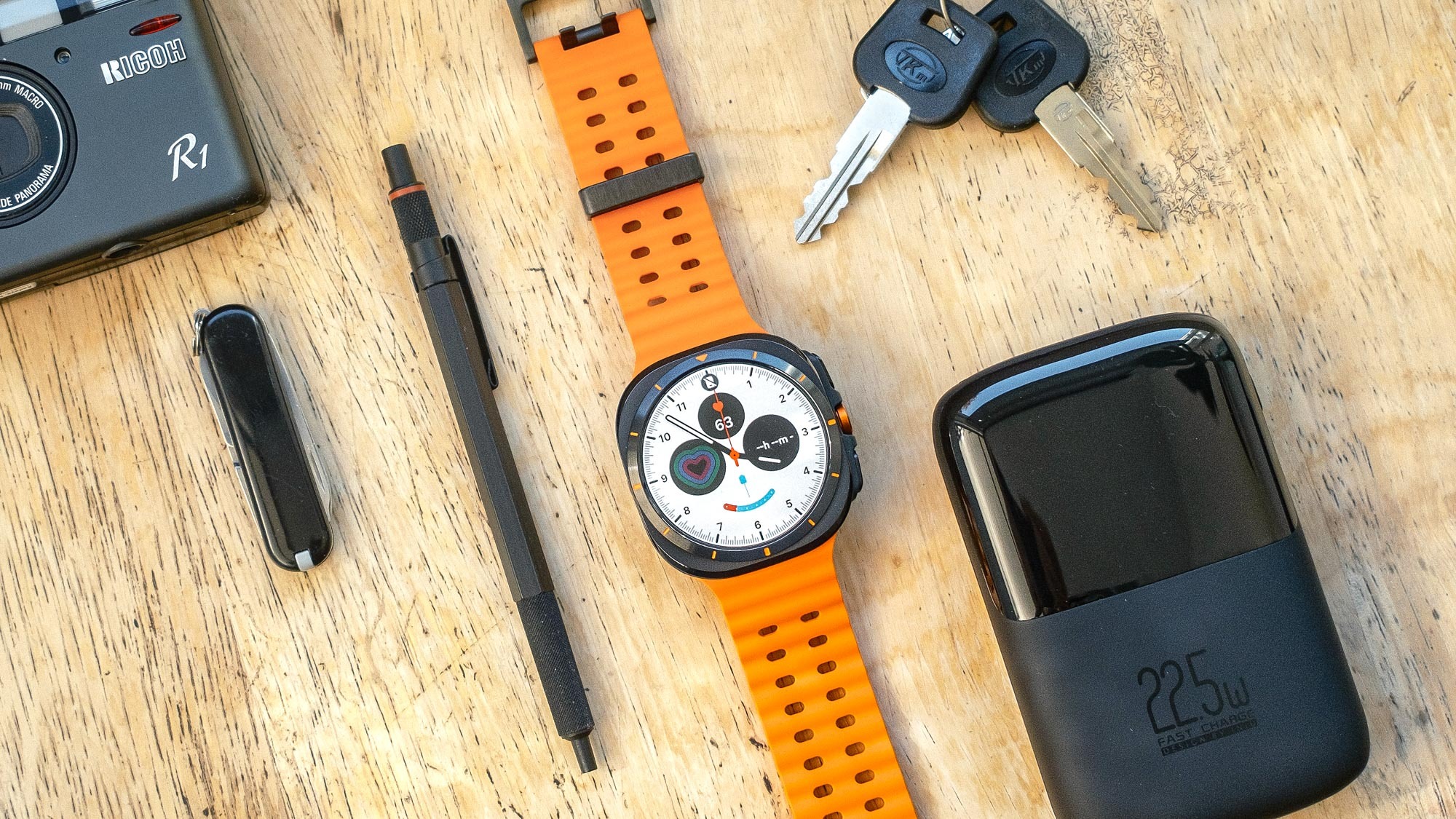 Samsung Galaxy Watch Ultra על רצועה כתומה יושב על שולחן עץ ליד אביזרים קטנים כמו מפתחות, עט ואולר
