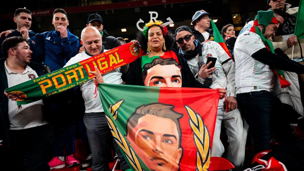 Woman seen wearing a CR7 - Cristiano Ronaldo shirt and holds flag with his portrait while surrounded by other soccer fans during a Portugal match.