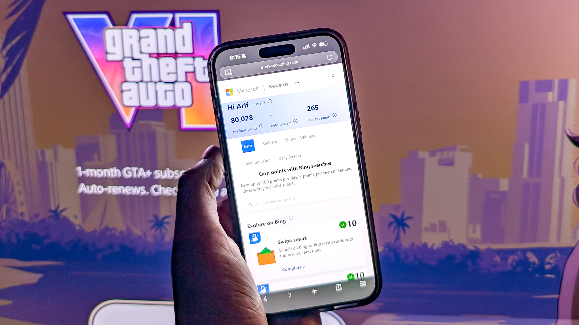 אדם שמחזיק טלפון עם יתרת Microsoft Rewards שלו מול דף ההזמנה המוקדמת של GTA 6