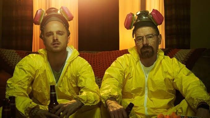 Actors Bryan Cranston and Aaron Paul, wearing yellow hazmat suits, sipping beer on sofa in a scene from Breaking Bad, the hit AMC crime series – here's where to watch and stream seasons 1-5