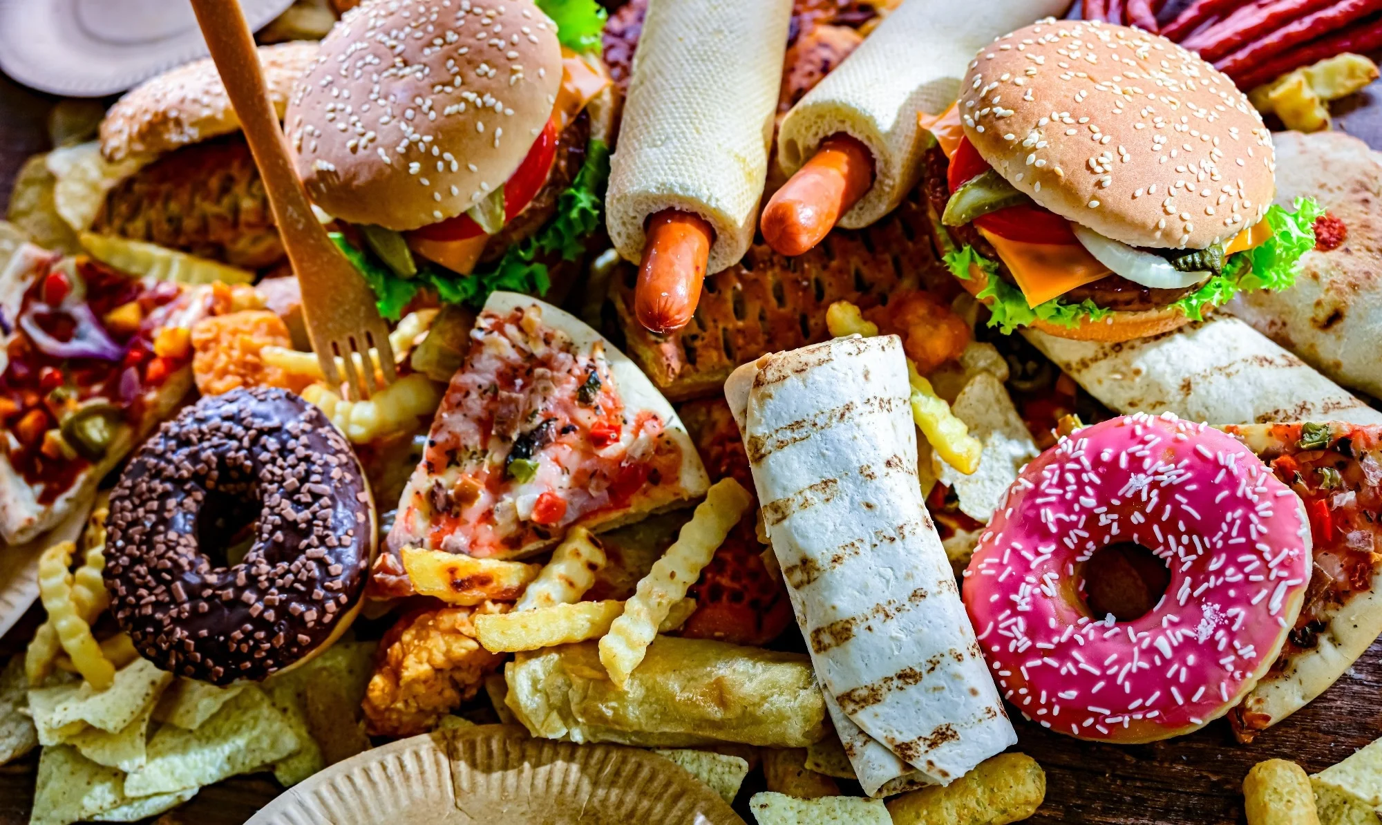 Assortment of ultraprocessed foods, including burgers, hot dogs, pizza, fries, and colorful doughnuts.
