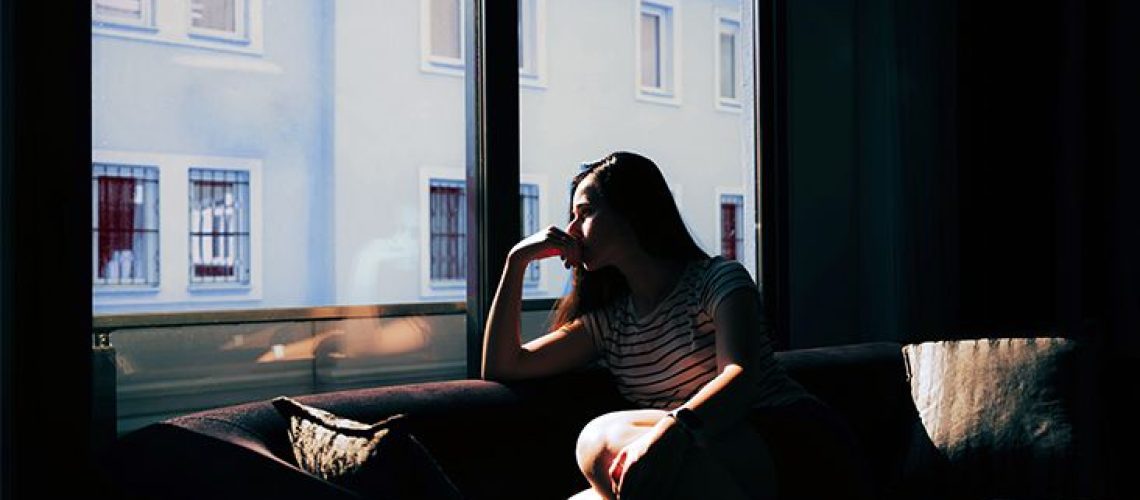 A woman looks out the window while sitting on a sofa in a dark room