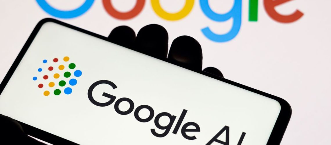 A Google AI logo is on a phone held in a hand, in front of a Google Logo