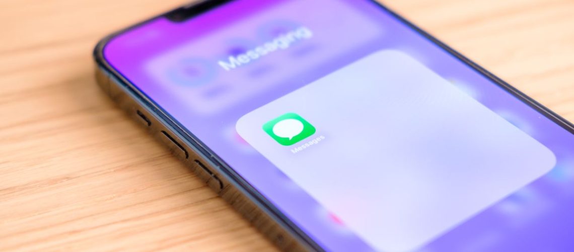 An image of an iPhone 13 Pro Max with a purple wallpaper. On screen is the iOS Messages app, opened within a Home Screen folder