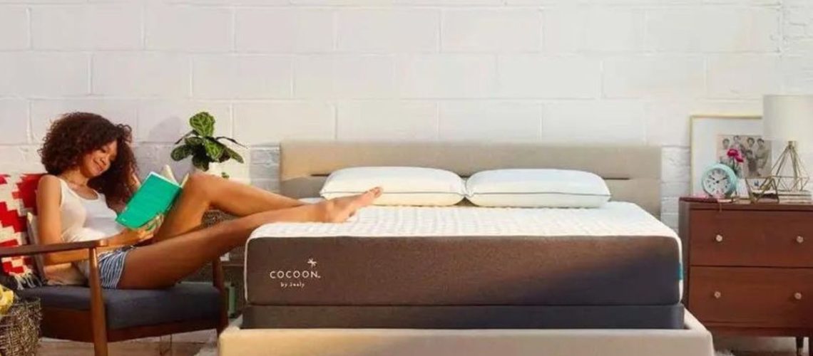 A woman wearing a white singlet an d denim shorts sits in a chair with her legs restiong on top of the Cocoon by Sealy Chill mattress