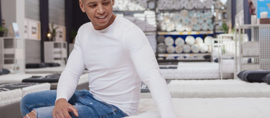 A man in a white long sleeve t-shirt places his hand on a mattress