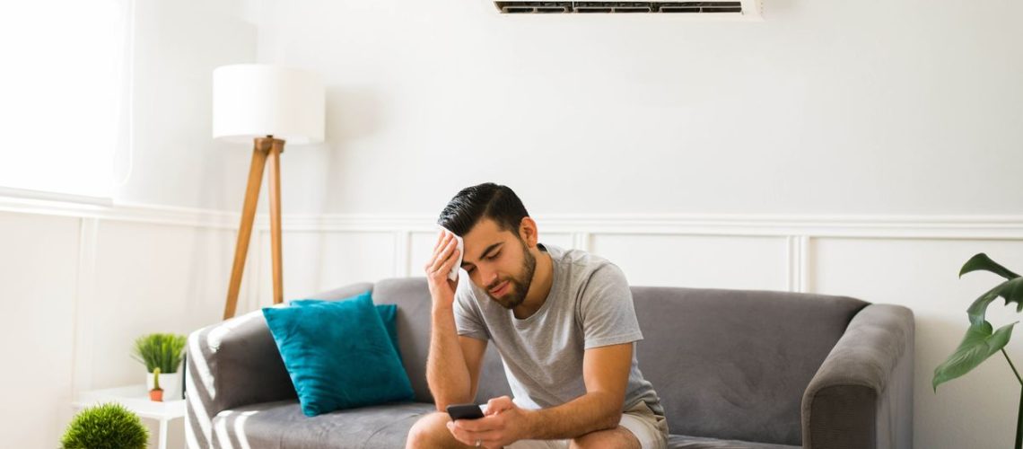 Man sweating on the sofa with AC unit behind him