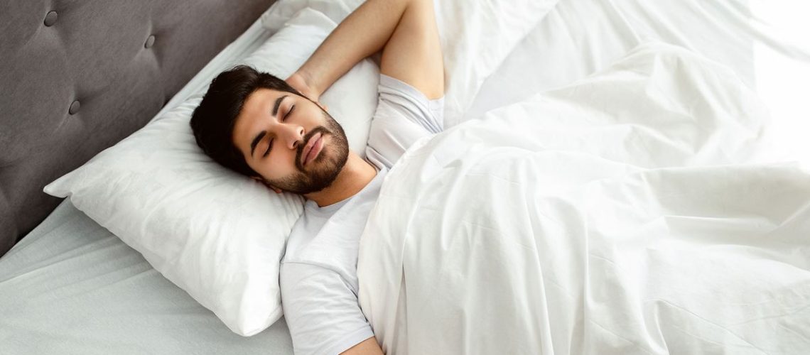 A man lies on his back in bed, sleeping happily in a comfortable position