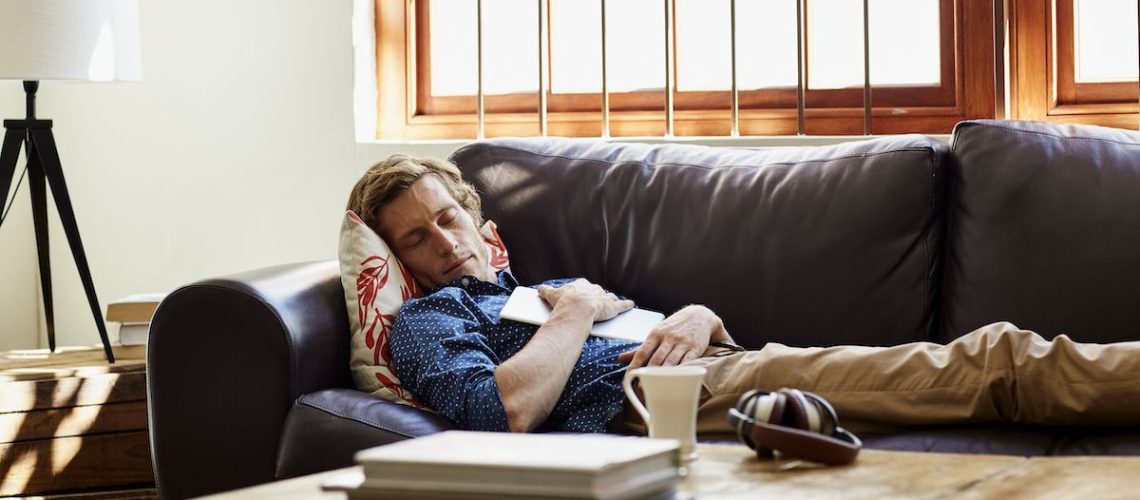 A blonde man wearing a blue shirt takes an afternoon nap on his brown leather couch during a summer heatwave