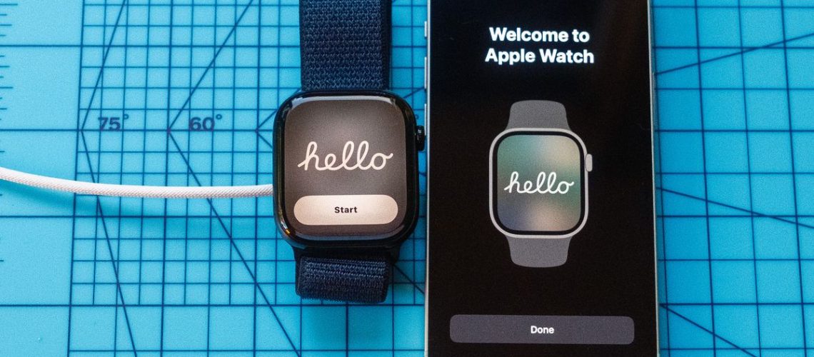 Close up of an Apple Watch 10 screen showing "Hello" next to an iPhone with the same message. Both are sitting on a light blue mat