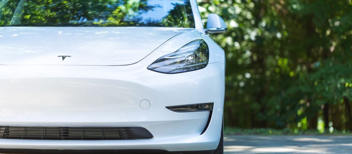An all electric Tesla Model 3 in white on cement road with trees in background on sunny day.