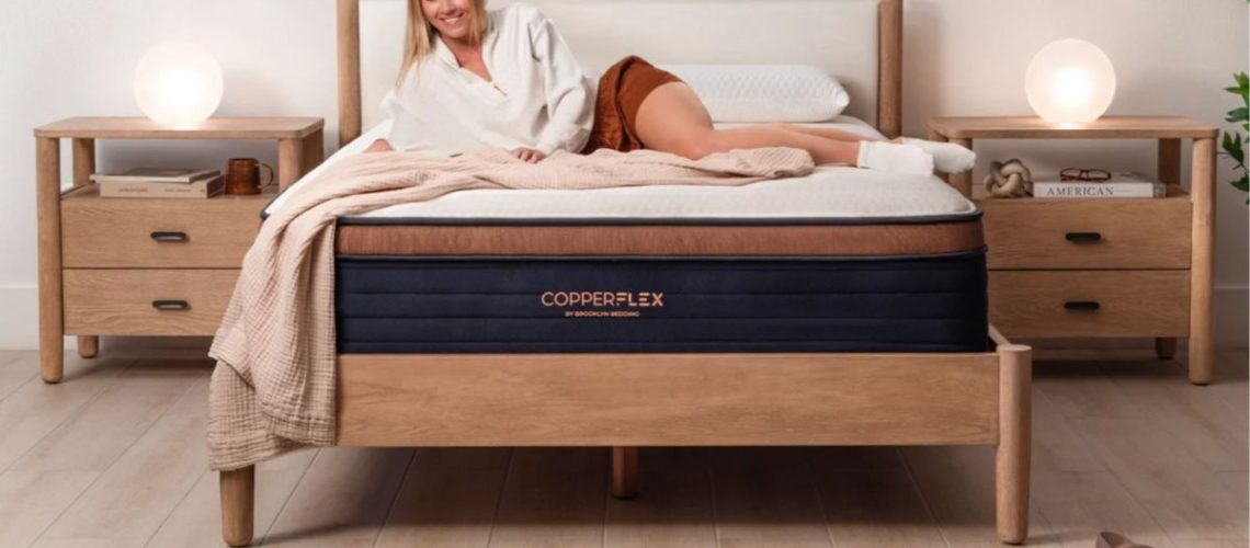 Blonde woman laying on Brooklyn Bedding CopperFlex mattress in bedroom with wooden floor, wooden bed base and two bedside tables