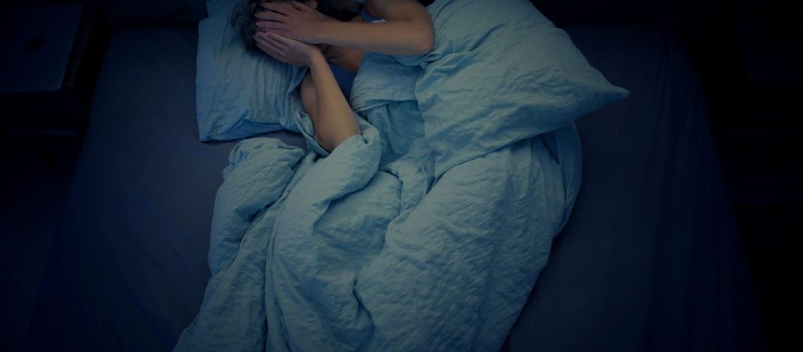 A woman lying in bed after waking from a night terror with her hands over her eyes, scared