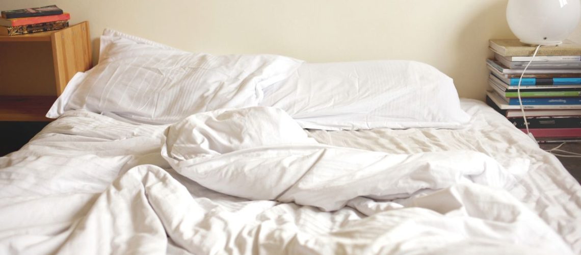 An unmade bed with a white duvet bunched up and white pillows, mattress shows slight body impressions