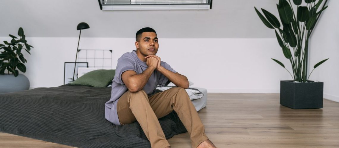 A man sits on a mattress on the floor placed on a hardwood floor so it sleeps cooler