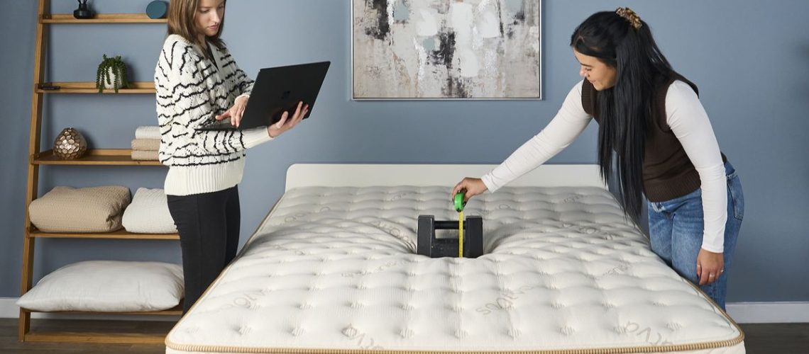 Two members of our review team photographed during a pressure relief test on the Saatva Classic mattress, with one typing notes into a laptop and the other measuring the amount of sinkage caused by a 56lb cast iron weight being dropped into the middle of the Saatva Classic mattress