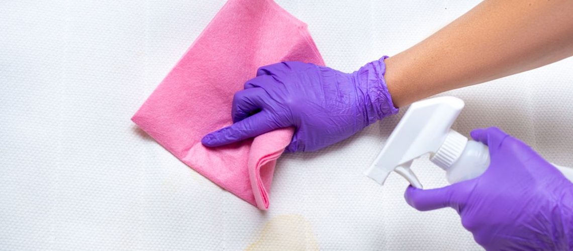 Image shows a person wearing purple disposable gloves using a pink cloth to clean yellow stains from the top of a white mattress