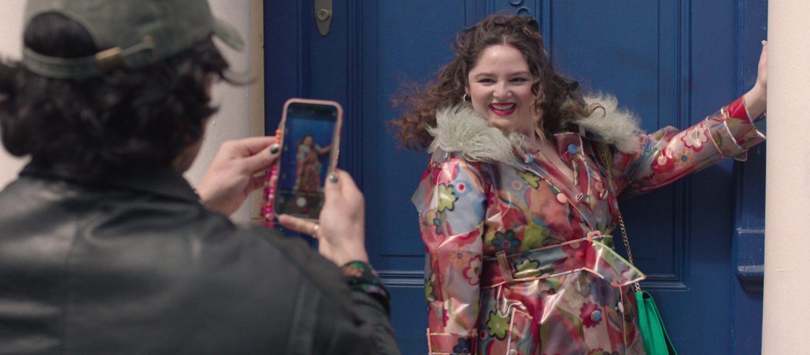 A man seen from behind takes a photo of Meg Stalter's character on his phone in a scene from Netflix's "Too Much"
