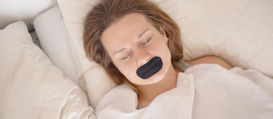 A woman lies asleep on her back in bed, her mouth covered with black mouth tape which she hopes will help her sleep better