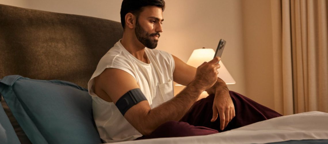 A man in white vest sitting up in bed checking phone with Garmin Index Sleep Monitor on right arm