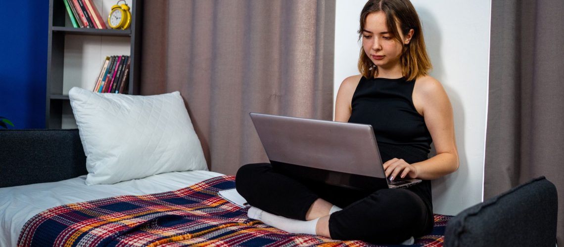 A young female college student with shoulder length brown hair looks at her laptop while sitting on her dorm bed.