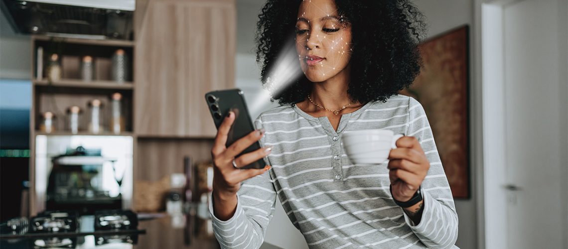Person, sat in a kitchen holding a mug, scanning their face with their smartphone