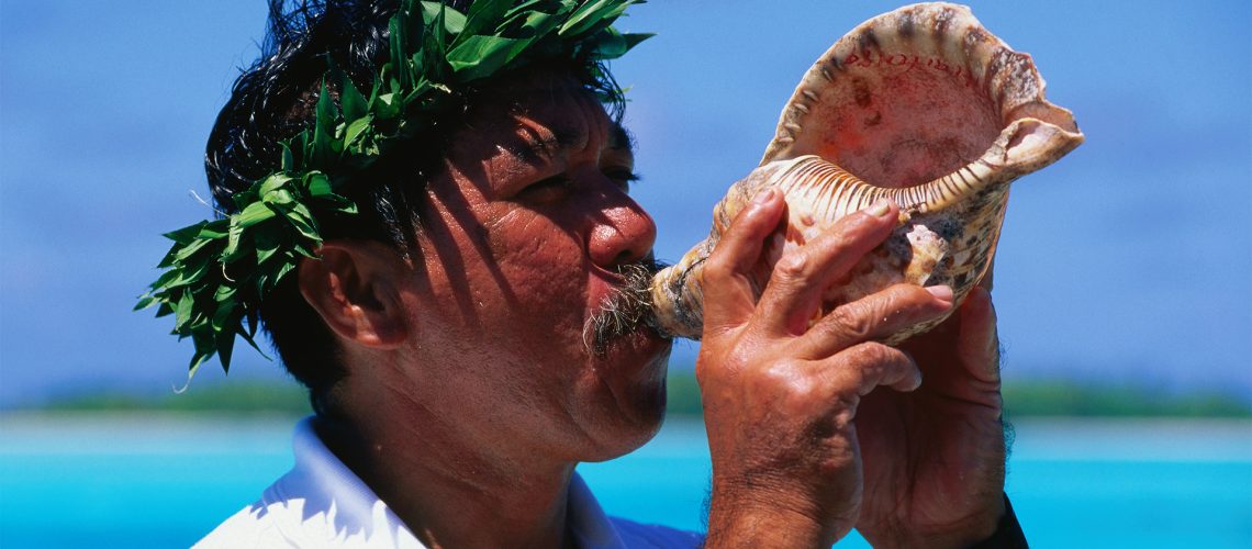 A man wearing a laurel holds a conch shell to his lips and blows into it, practicing the ancient ritual of shankh breathing