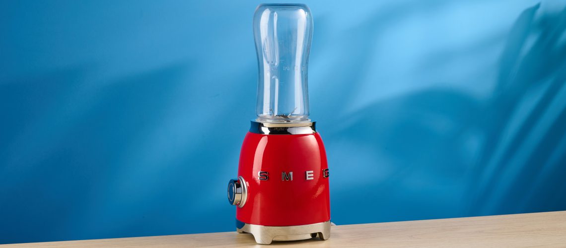 Smeg Personal Blender in the red colorways photographed in front of a blue background.