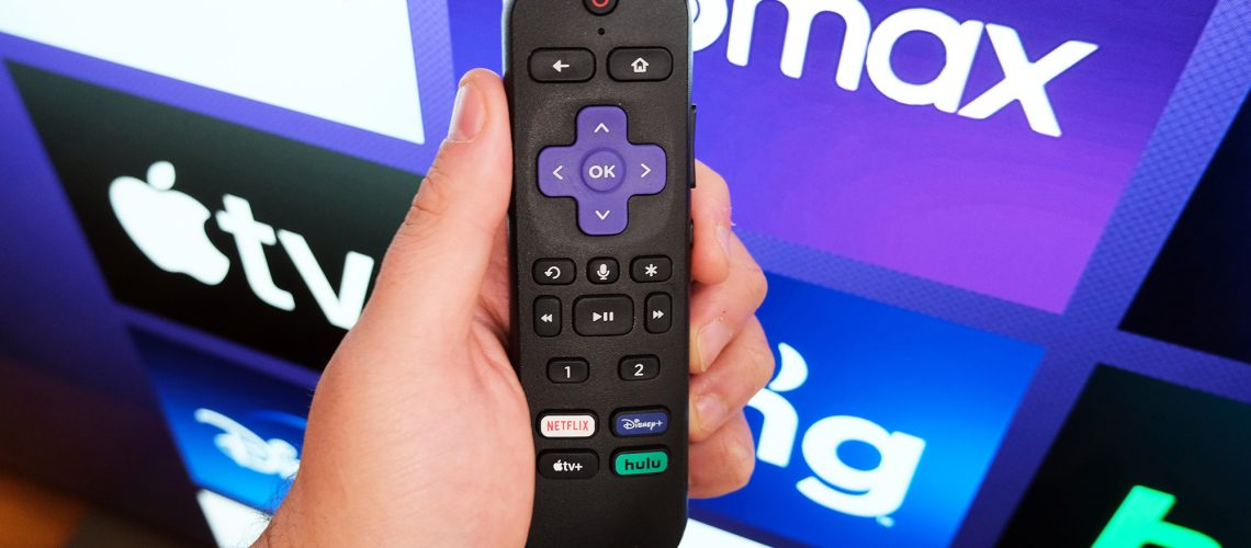 A Roku remote in hand in front of a TV with the Roku home page.