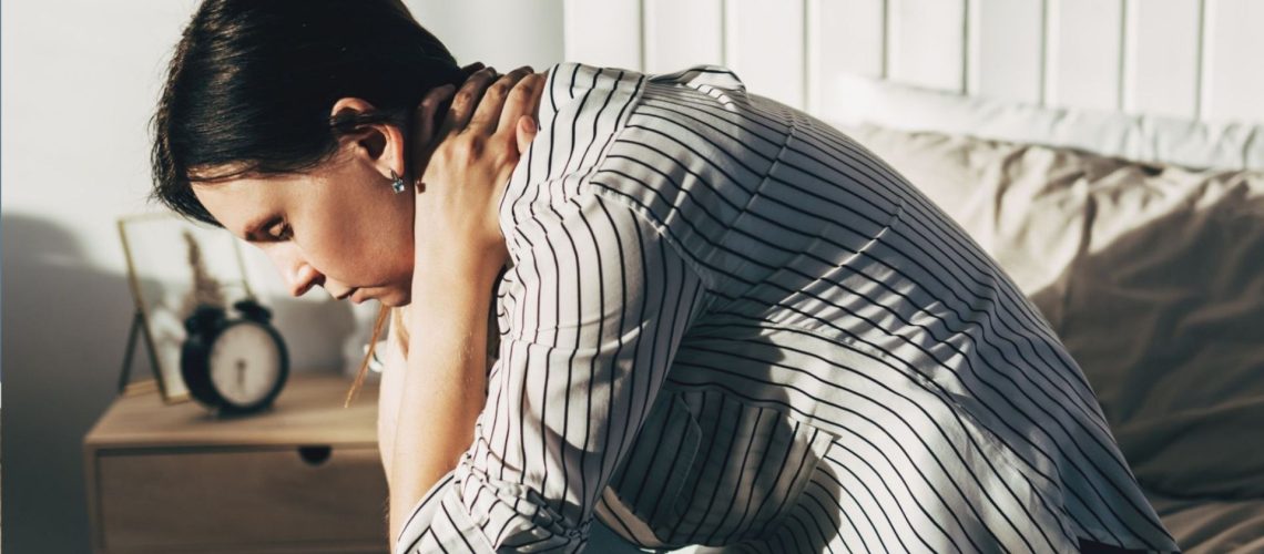 The image shows a dark haired woman wearing a stripy shirt sitting on the edge of a bed and holding her head in her hands