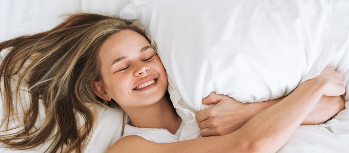 A woman lays in bed, smiling with her eyes closed, while holding her pillow.