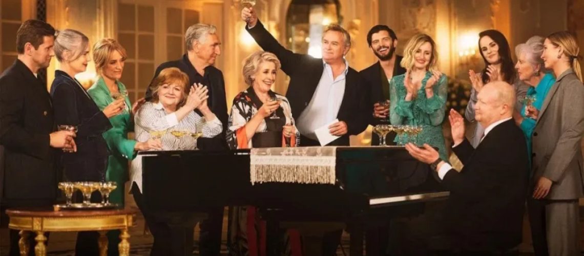The cast of "Downton Abbey" standing around a grand piano and celebrating in a promotional shot for "Downton Abbey Celebrates the Grand Finale"
