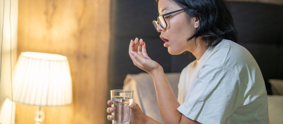 A woman with black hair wearing glasses and holding a glass of water sits on the edge of her bed and prepares to take a multivitamin supplement at night.