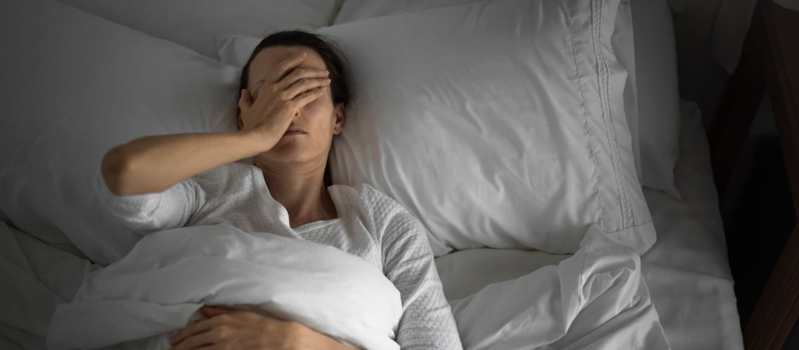 A woman lies on her back in a bed that has white bedding, covering her face with one hand.