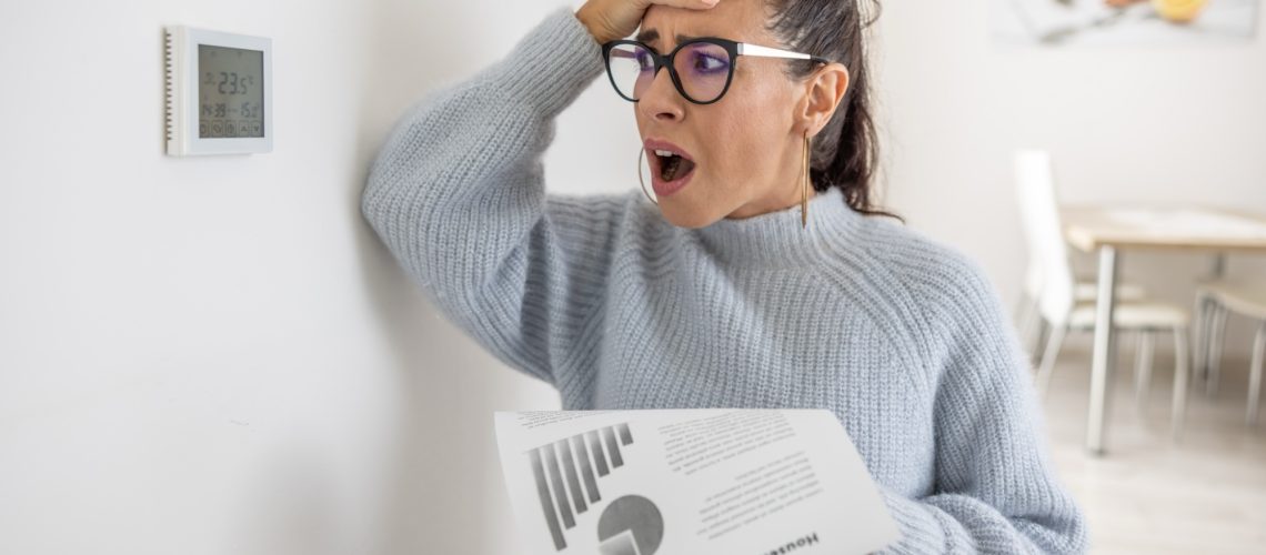 woman holding energy bill next to thermostat looking upset