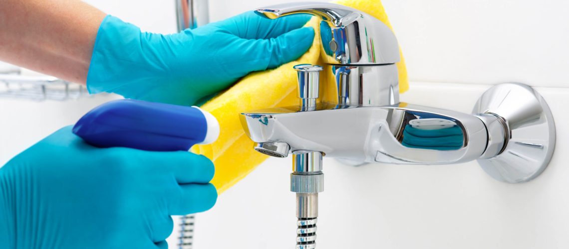 Someone cleaning a bathroom faucet with a spray bottle and cloth while wearing gloves