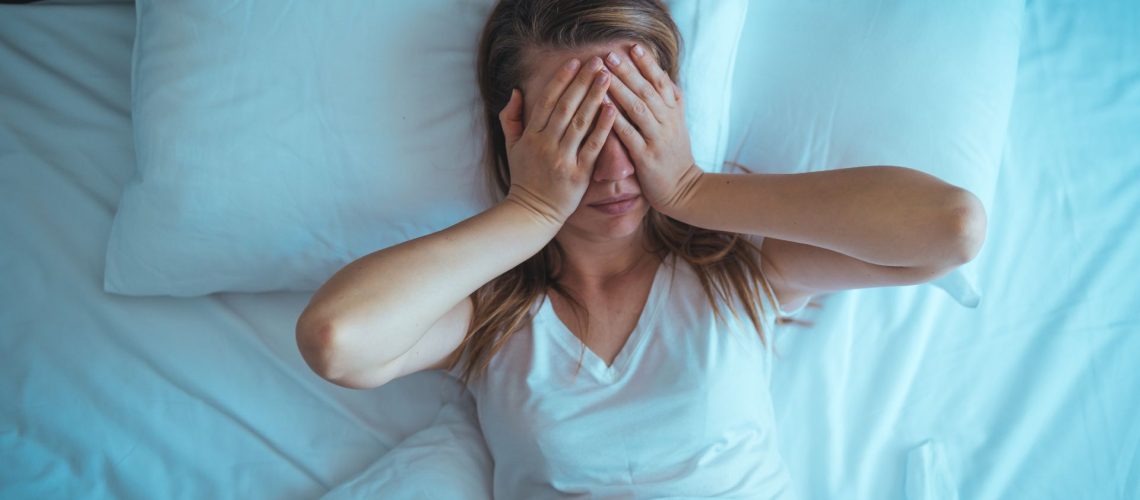 A woman lies in bed covering her eyes with her hands as she struggles to sleep.