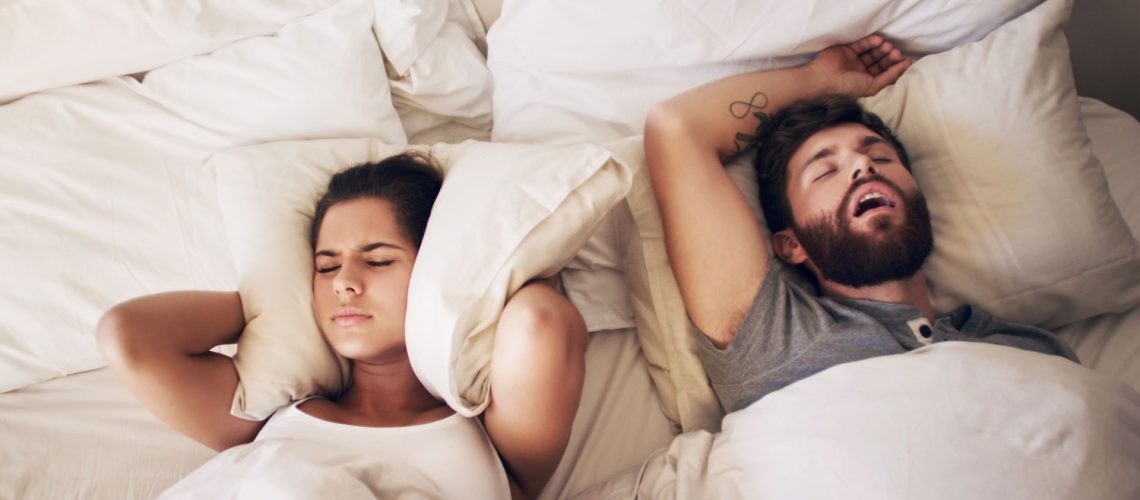 A man snores while asleep in bed as a woman next to him covers her ears with a pillow to block out the noise.