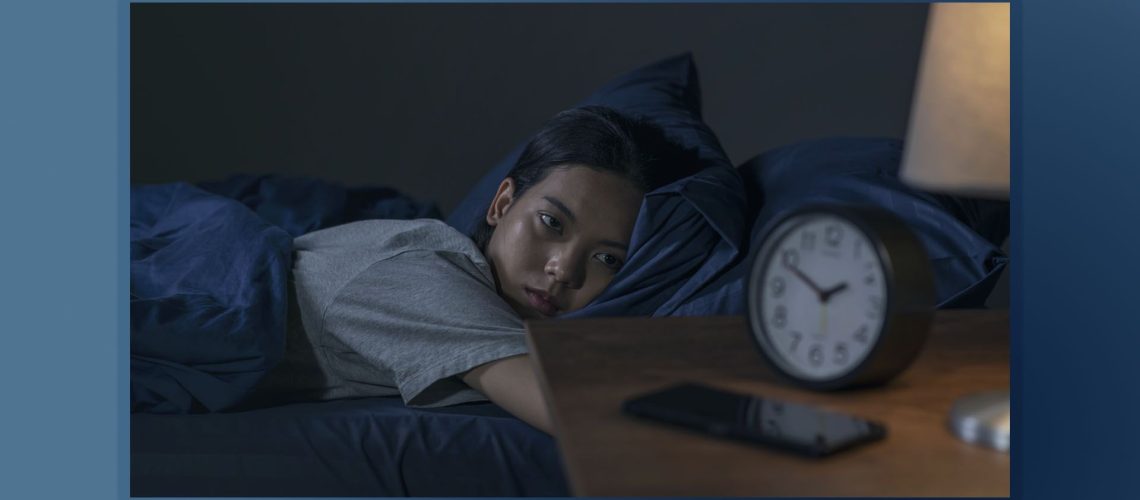 A woman lies in bed in the middle of the night unable to sleep, watching her alarm clock which is placed on a nightstand in the foreground of the image