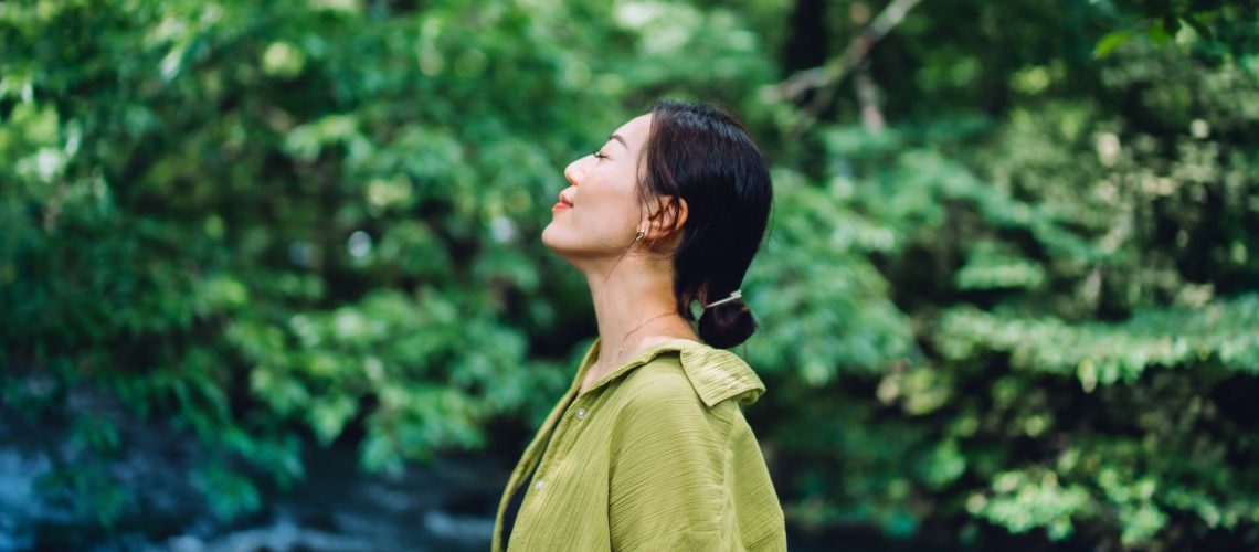 Woman in green blouse standing in nature with eyes closed smiling