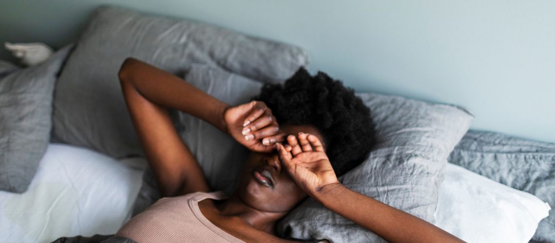 A woman lies in bed in the morning rubbing her eyes as she lays on gray and white bedding.